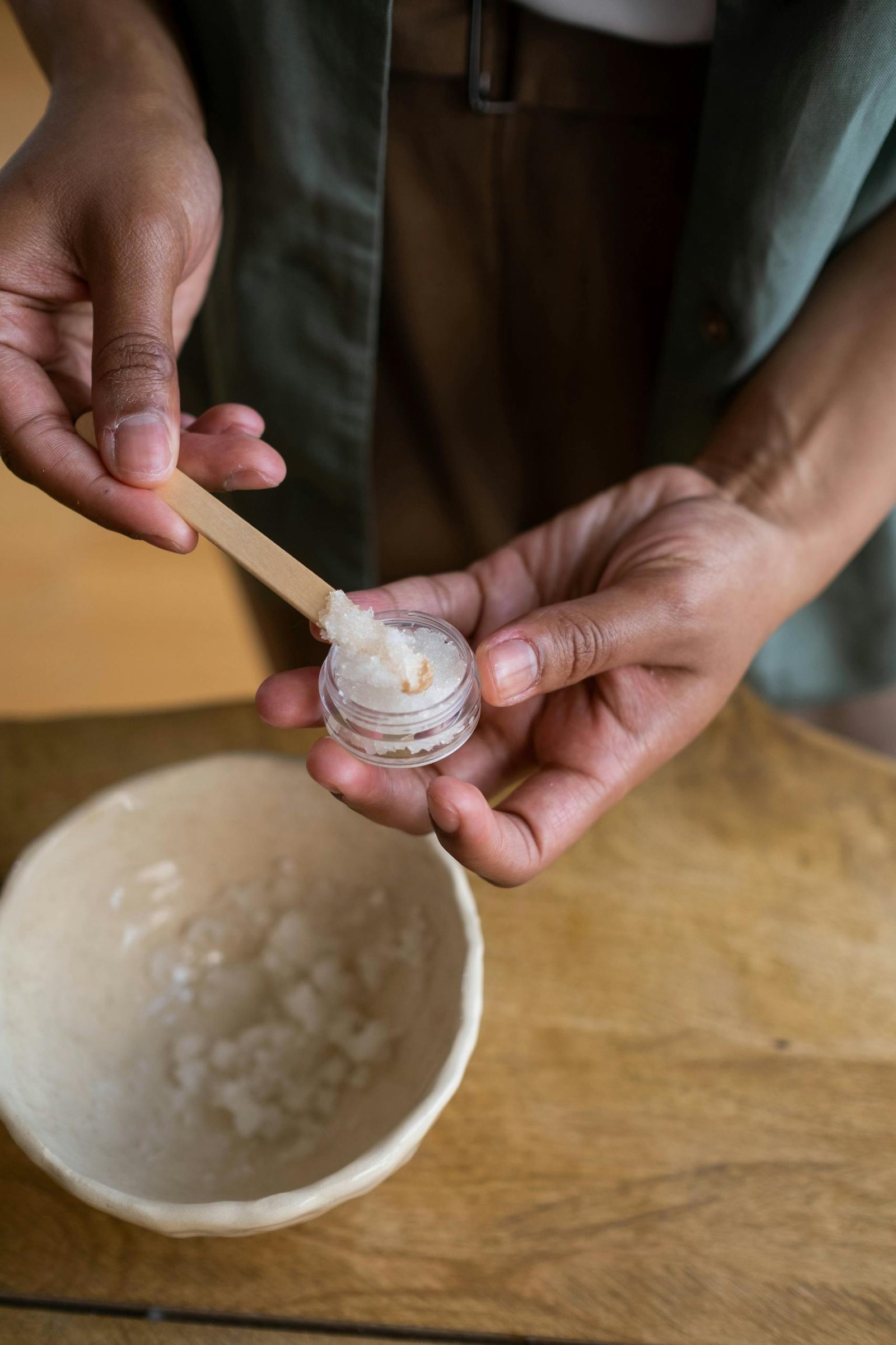 A person using a wooden stick to carefully transfer ointment from a bowl into a small container.