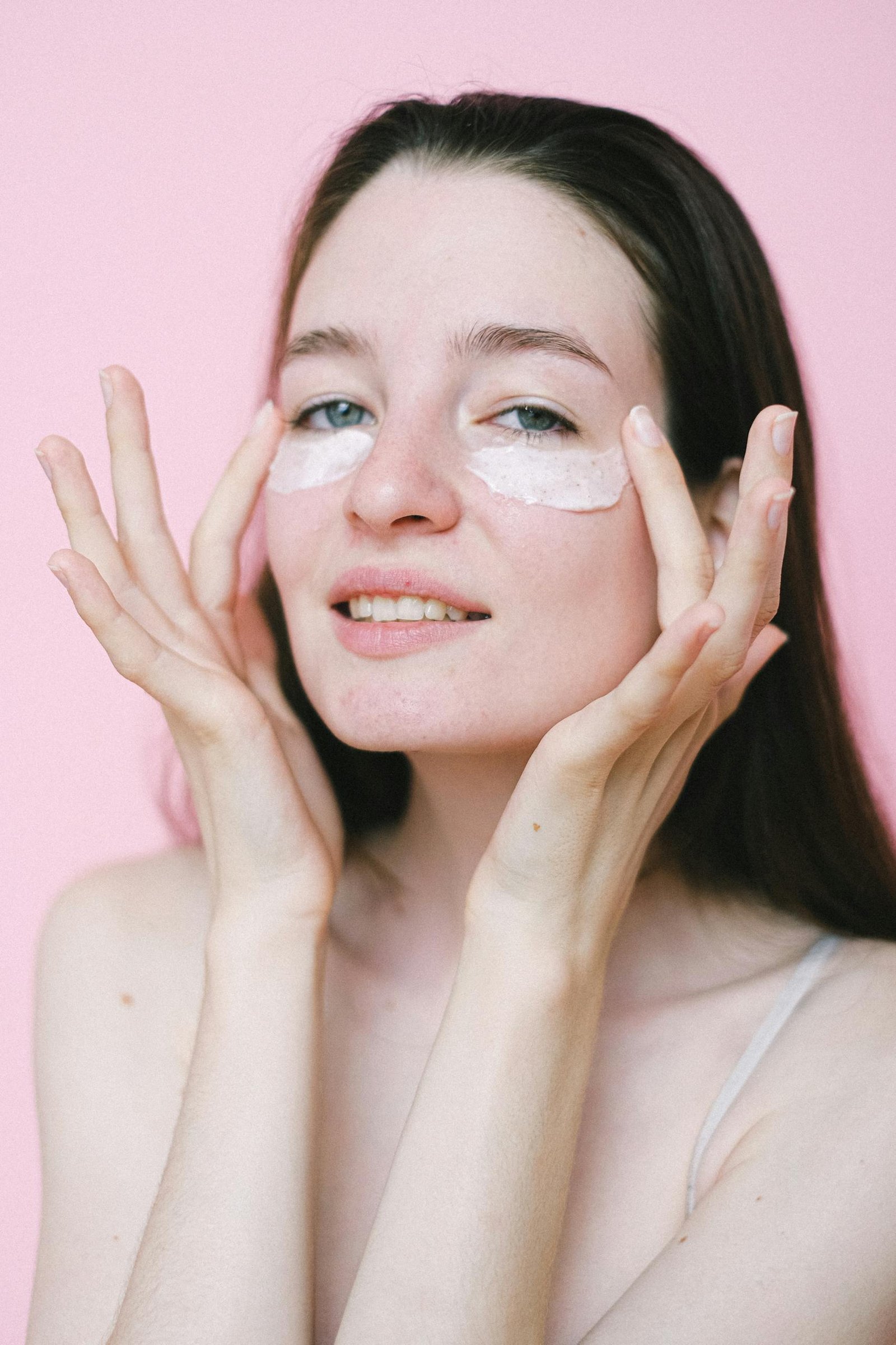 Portrait of a young woman applying under-eye cream in a studio setting with a pink background.