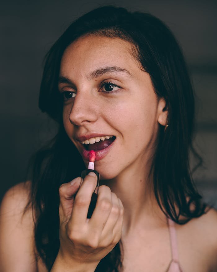 A young woman applies pink lipstick indoors, showcasing natural beauty.